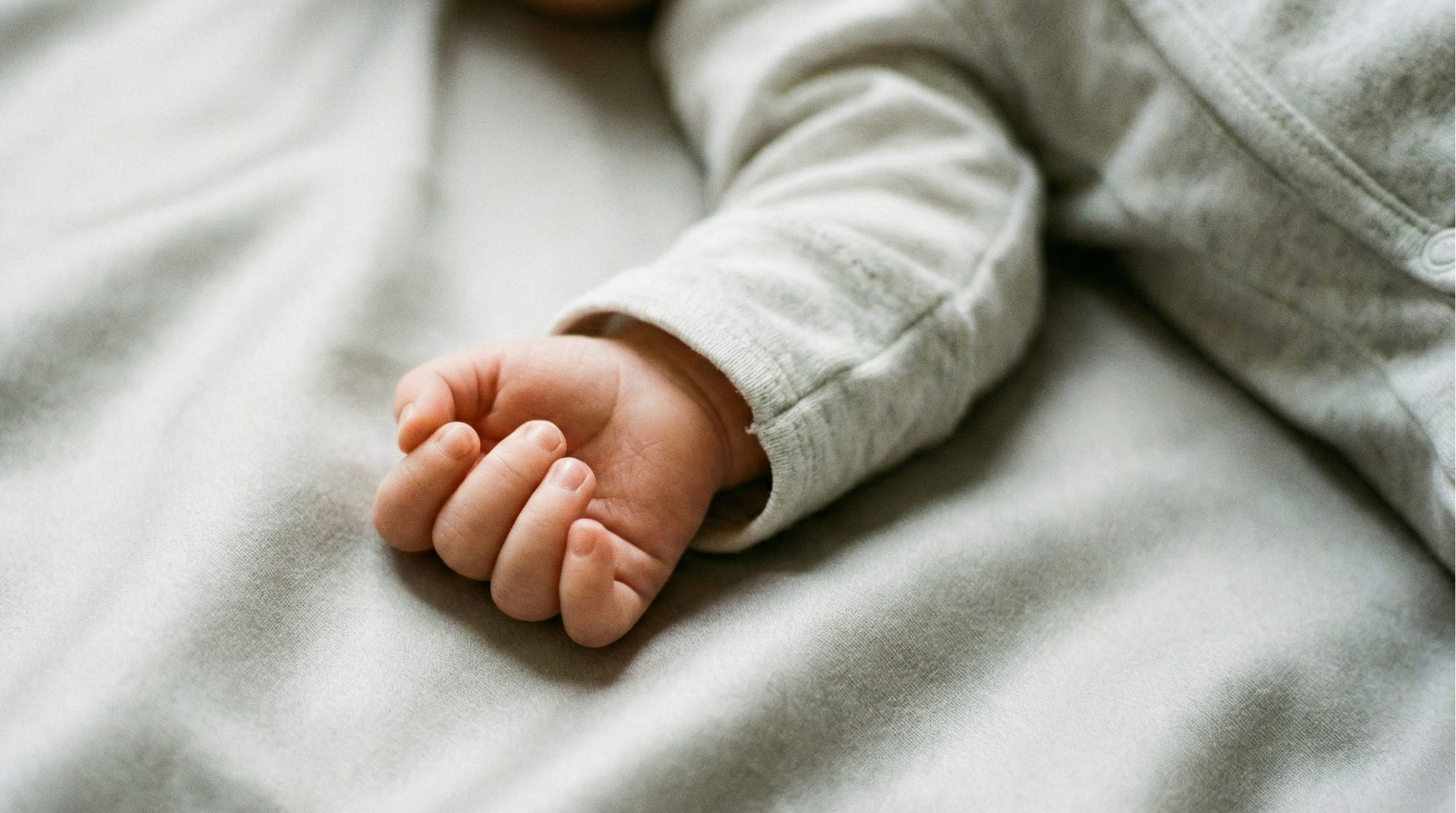 Nahaufnahme einer zarten Babyhand und des Unterarms, die auf weicher, hellgrauer Tencel-Satin-Bettwäsche ruhen, wodurch die seidige Textur und Hautfreundlichkeit des Materials verdeutlicht wird.