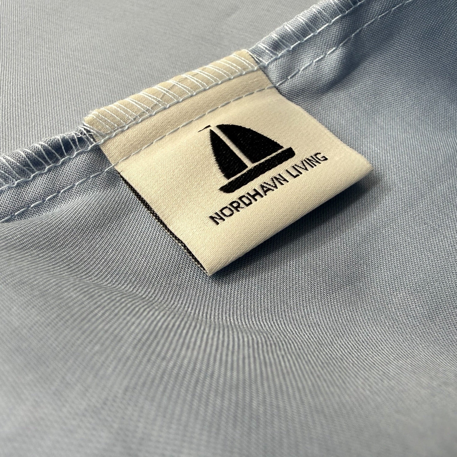 Nordhavn Living Aurora Satin Bettwäsche in Kühlem Graublau (Frost Haze): Detailansicht des eingenähten Marken-Labels mit Segelboot-Logo auf dem fein schimmernden Tencel-Stoff.