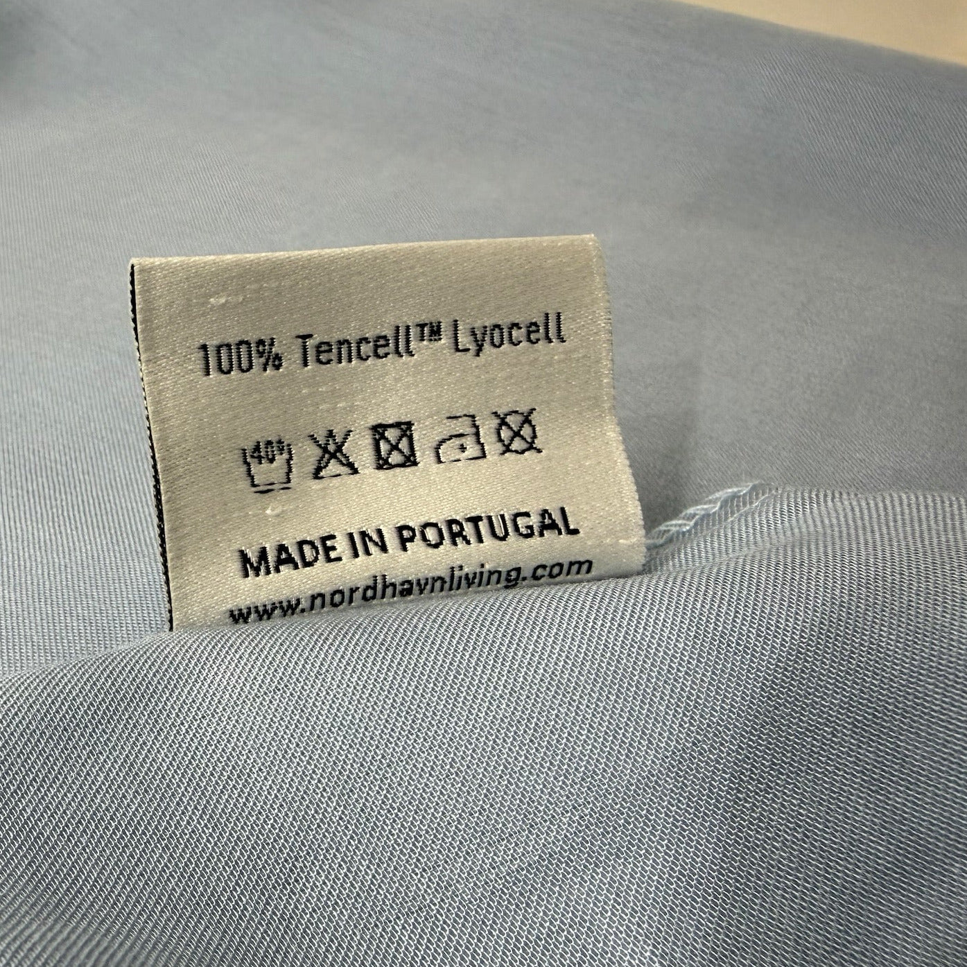 Nordhavn Living Aurora Satin Bettwäsche in Kühlem Graublau (Frost Haze): Detailansicht des Einnähetiketts mit Pflegehinweisen, der Materialangabe 100% Tencel™ Lyocell und der Herkunft Made in Portugal.
