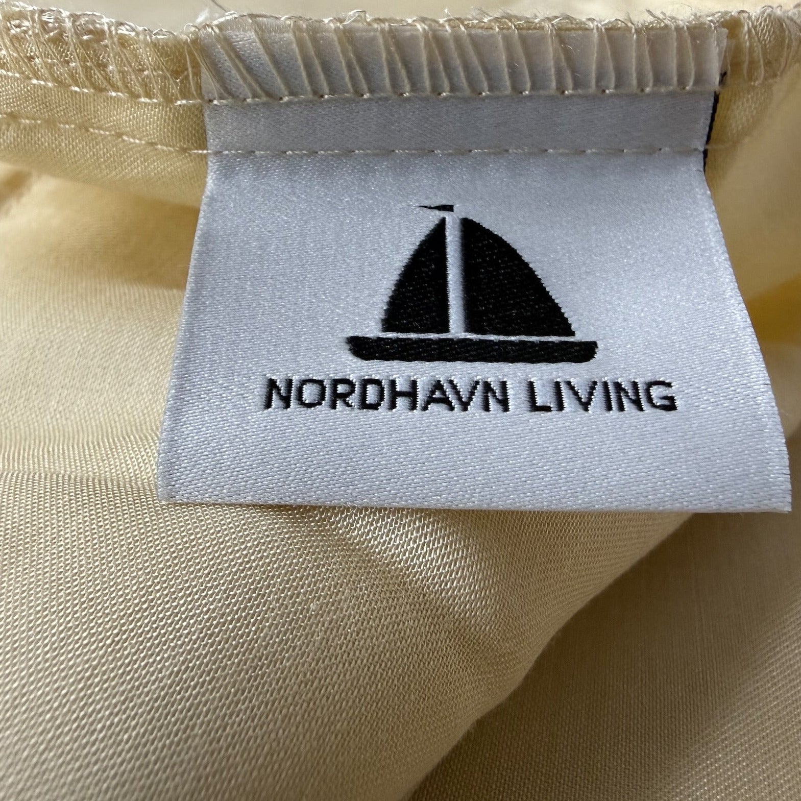 Nordhavn Living Aurora Satin Bettwäsche in Hellem Greige (Harvest Dust): Detailaufnahme des eingenähten Marken-Labels mit dem schwarzen Segelboot-Logo auf dem cremigen, seidigen Satin-Gewebe.