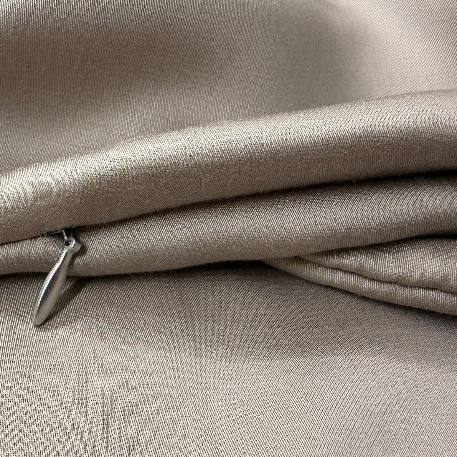 Nordhavn Living Aurora Satin Bettwäsche in Warmem Lehmgrau (Still Clay): Detailaufnahme der weich fallenden Stoffbahnen mit Fokus auf den hochwertigen, silbernen Metall-Zipper des verdeckten Reißverschlusses.