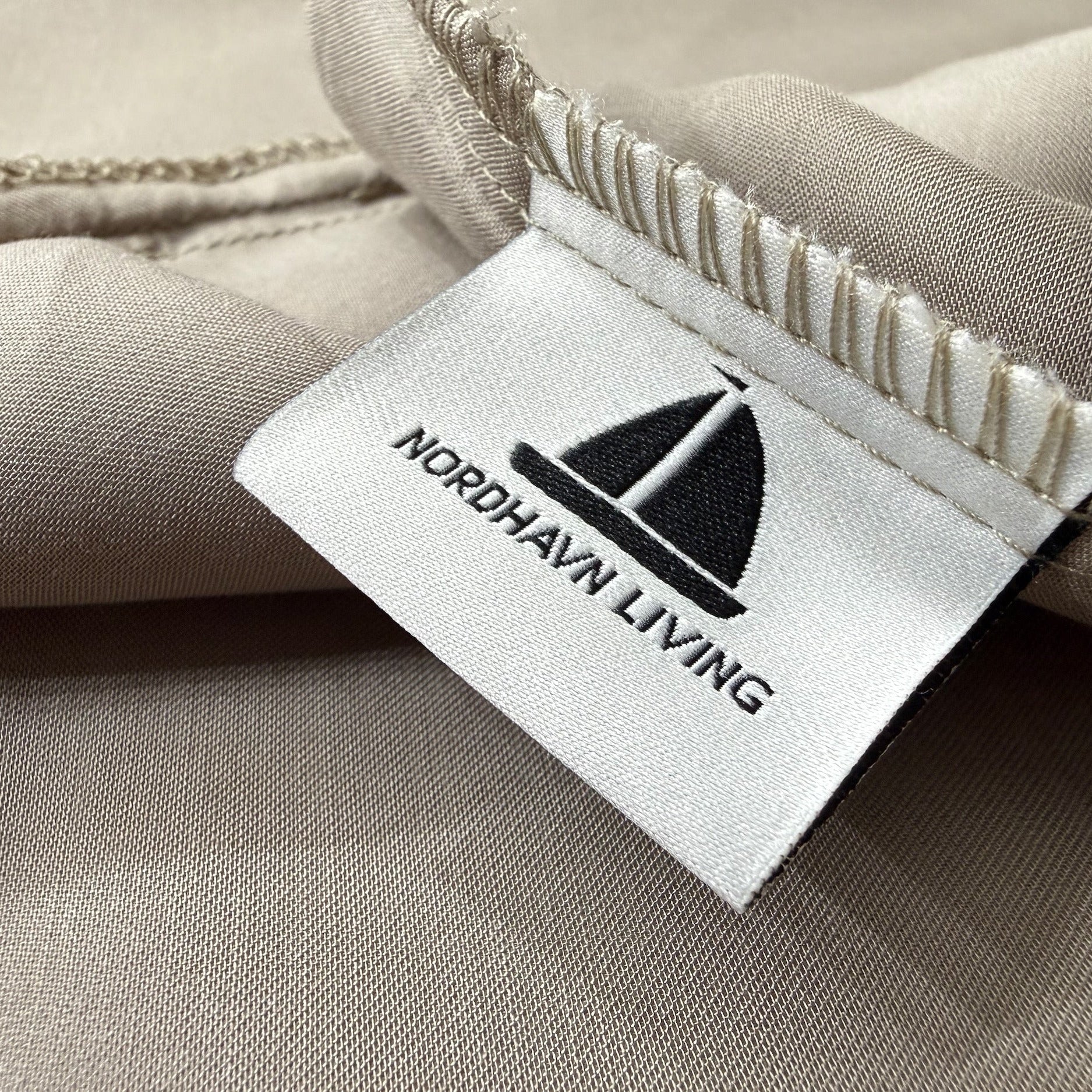 Nordhavn Living Aurora Satin Bettwäsche in Warmem Lehmgrau (Still Clay): Detailaufnahme des eingenähten Marken-Labels mit Segelboot-Logo auf dem natürlich schimmernden, erdigen Tencel-Stoff.