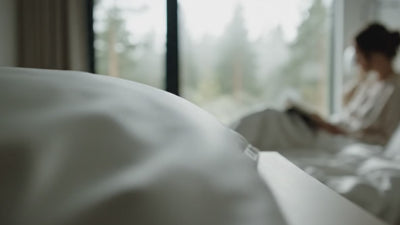 Minimalistisches Luxus-Schlafzimmer mit bodentiefen Fenstern und Waldblick; im Vordergrund weiße TENCEL™ Lyocell Satin Bettwäsche mit softem Glanz, ein Model liest entspannt auf dem Bett.